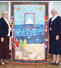 Two older ladies stand by a large quilt (with a seafront design, including beach, sea and beach huts).