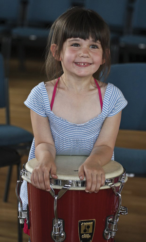 A small girl grinning at the camera and leaning on a samba drum