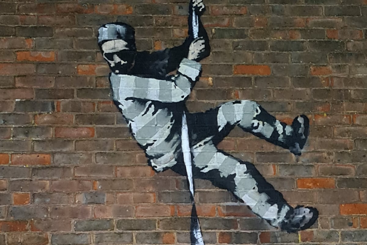 Black and white stencil graffiti of a prisoner escaping down a wall