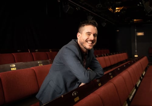 Michael Longhurst is a man. He has brown hair shaved on the sides. He has stubble and has a moustache. He is wearing a grey blazer. He is stood in the middle of a row of chairs in the auditorium of Donmar Warehouse.