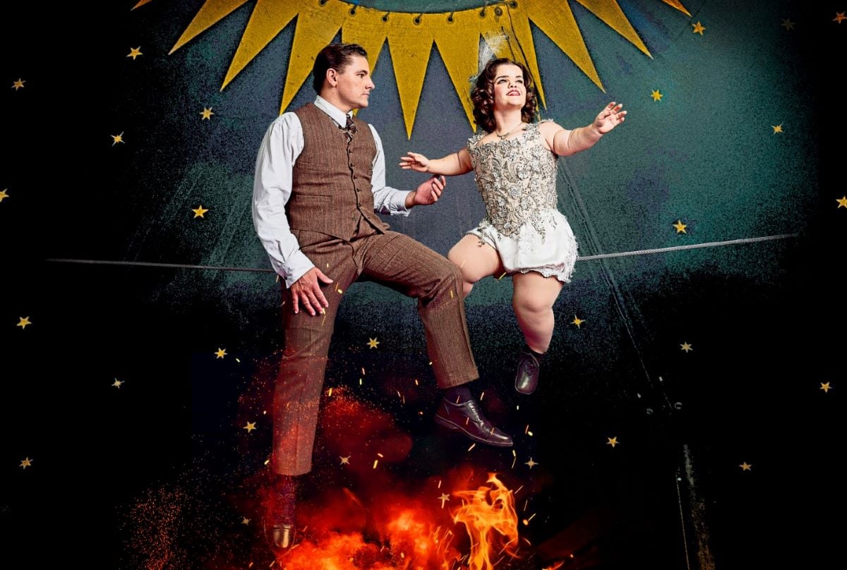 The image is two performers on a raised tight wire in a dark circus tent decorated with gold stars and bunting. On the right is Krista, a young white woman of short stature with short brown hair looking into the distance. On the left is Gerhard, a young white man with short brown hair, who gazes intently at the other performer, Krista, as he reaches his hand to touch her. Both characters’ costumes are in the style of the 1930s. Hers is a shiny silver beaded corset with white silk shorts, tights and black sh