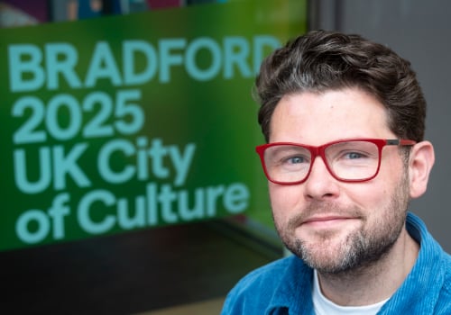 Description of Will White: A white man with brown hair, green eyes, and a short beard and moustache, wearing red glasses and a bright blue corduroy shirt. He stands or sits in front of a green sign that reads 'Bradford 2025 UK City of Culture'.