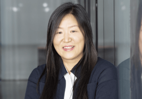 Sook-Kyung Lee, incoming Director, Whitworth Gallery. She is a Korean-born woman with dark, long hair. She wears dark clothing, and is positioned leaning against a glass wall, folding her arms.