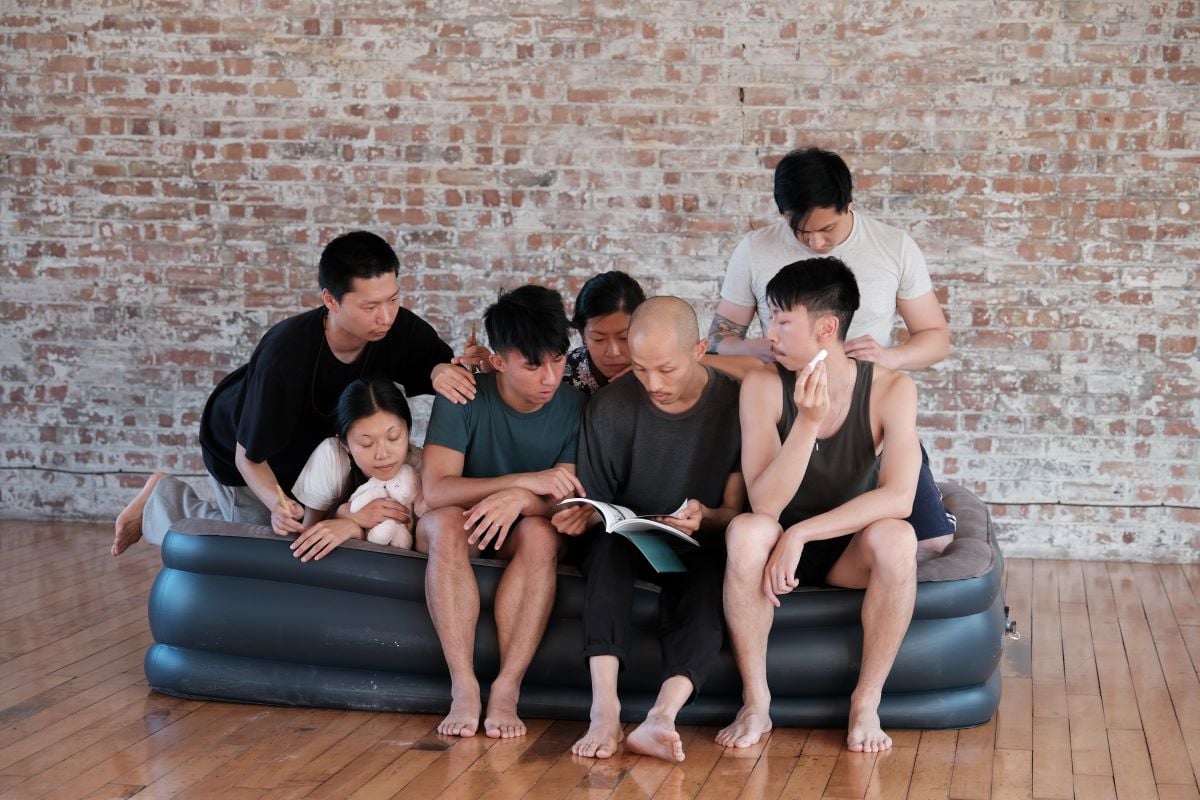 Chisenhale Dance Space August Residencies 2022, Ghost and John. People sat together on a blow-up mattress in a dance studio. They are looking from a book the person in the middle is reading from.