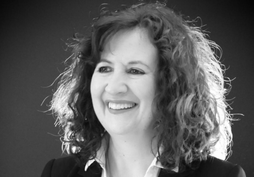 Black and white headshot of Jane Cordell. She has curly, shoulder-length hair, and wears a white shirt and black blazer. Smiling, she looks to the right away from the camera.