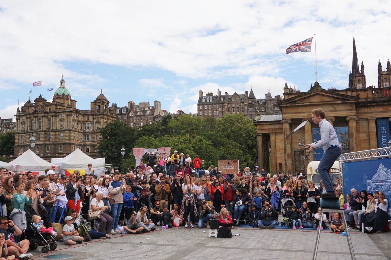 Edinburgh, Scotland - August 8, 2018: This is one of the many performance areas set up during the Fringe Festival where street performers and performance groups have time slots for entertaining the passing crowds. A juggler with some rather sharp implements has attracted a large crowd in the plaza beside the art galleries on the corner of Princes St and the Mound. In the background is the skyline of the historic High St / Royal Mile.