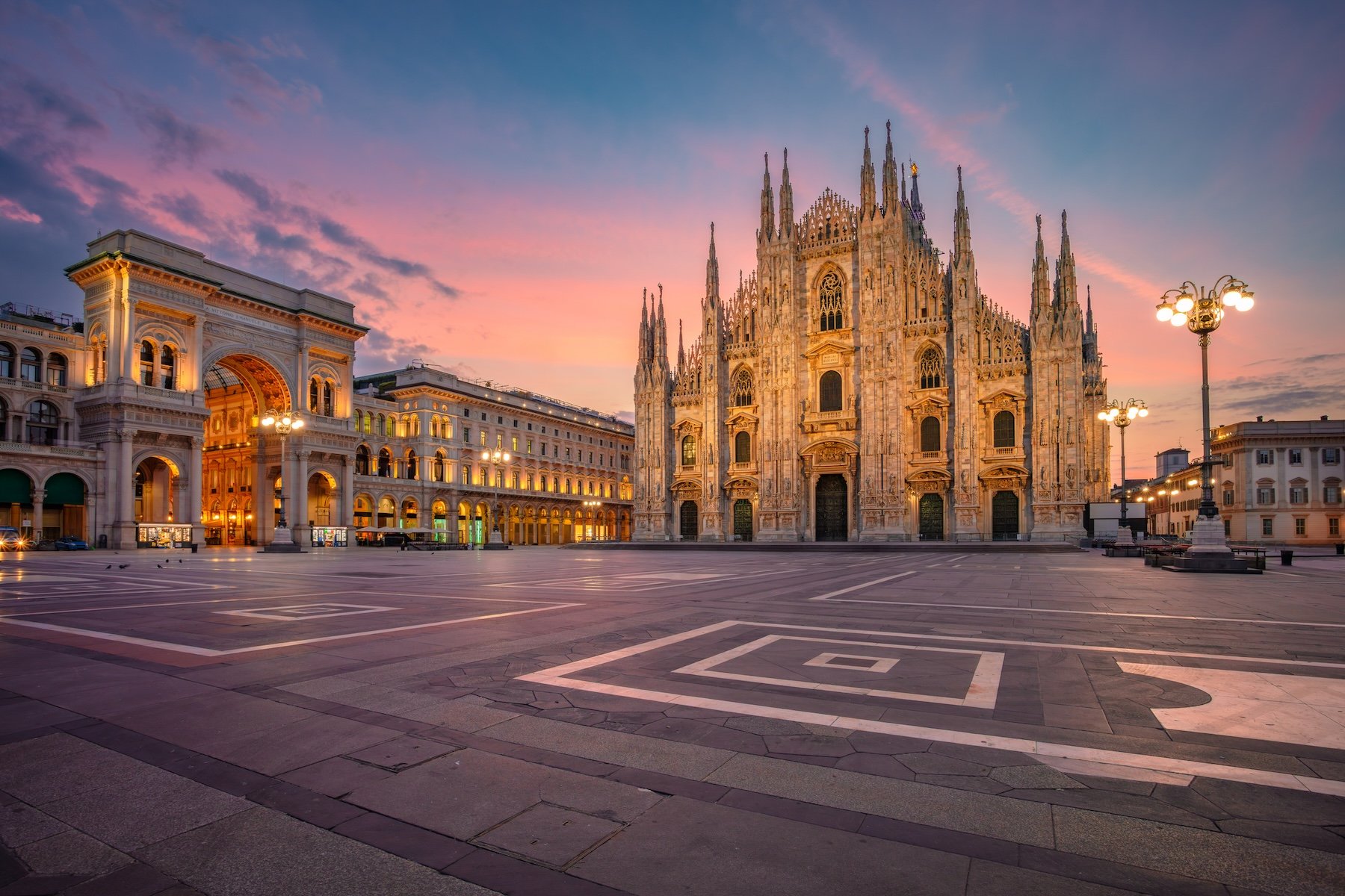 Milan Cathedral, or Duomo di Milano, stands prominently at sunrise in Milan, Italy. The Gothic architecture is illuminated by soft morning light, highlighting its intricate spires and façade. Nearby, Galleria Vittorio Emanuele II, an arched, glass-vaulted arcade, is visible. The surrounding piazza is spacious and appears empty, with the sky painted in pastel hues of pink and blue. Historic street lamps add to the serene ambiance of this iconic cityscape.