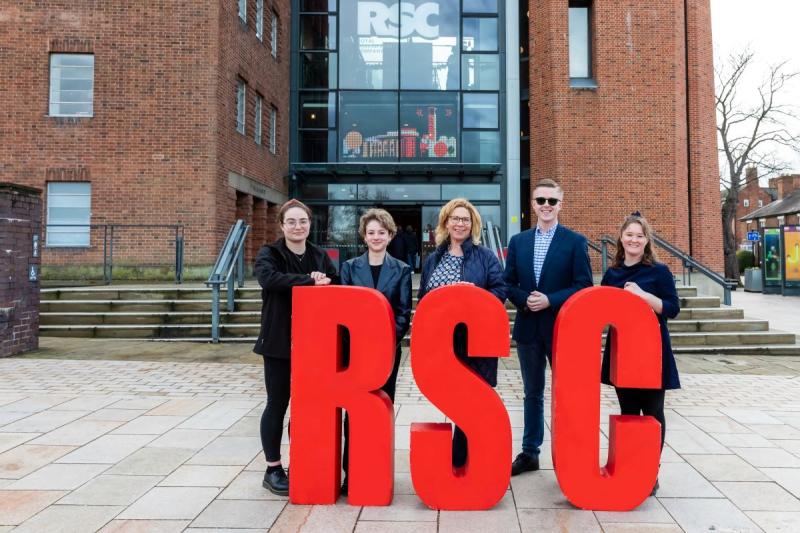 Four RSC apprentices stood in front of Royal Shakespeare Company's building. They are stood directly behind three big red letters that reads 'RSC'. They are looking and smiling at the camera.