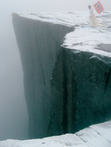 A cloaked man holds a Norwegian flag on the edge of a icy cliff
