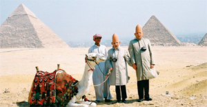 Men with Cone heads stand in front of the Pyramids, with a camel and a man in a turban