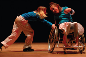 A lady puses another lady's wheelchair as part of a performance on stage