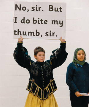 A boy dressed in Shakespearean costume holds up a sign reading 'No, sir. But I do bite my thumb, sir.