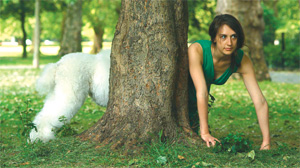 A girl on all 'all fours' appears to stick out from behind a tree, instead of her back end being visible at the other side of the tree, we can see the rear end of a poodle.