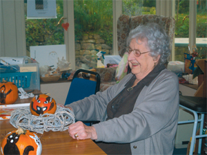 An older lady sits at a table, on teh table is a painted orange ball and some rope, perhaps a craft project for Halloween.