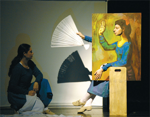 A lady kneels on stage, at the foot of a portrait, which holds a fan (the arm holding the fan, and the face on the painting are real)