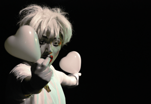A masked performer on stage holds two plastic hearts.