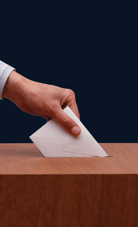 A hand puts a envelope into a ballot box (as if casting a vote)