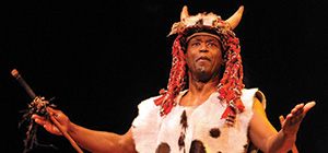 A man on stage in tribal costume (including a hat with horns)