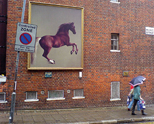 A painting of a horse (in a gold frame, reminiscent of one you may find in a stately home) hung on a red brick building's wall outside.
