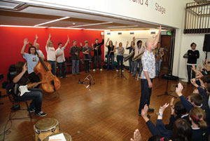 A group of musicians stand in front of a hall of children with their hands up.