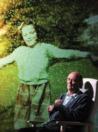Actor, sitting in an arm chair, on stage with an image of a young child projected behind on a screen