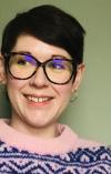 Hannah Slimmon. She has short black hair and wears tortoise shell glasses and a colourful purple/pink jumper. She is photographed smiling against a green wall.