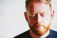 has ginger hair and beard and looks downwards with a subtle frown. Positioned against a white backdrop, he is captured to the side of the frame. He wears a dark blue t-shirt.