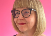 Annabel Turpin, incoming Chief Executive of Storyhouse (Chester). She has a short, blonde bob hairstyle with a full fringe. She wears black, large glasses and red lipstick. She is captured smiling and glancing sideways, with a bright pink wall in the background.
