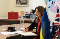 Culture Secretary Lucy Frazer sits at a desk, she is speaking and writing notes. UK and Ukrainian flags are in the foreground