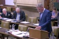 MPs listen to DCMS Minister Stuart Andrew during the debate. Andrew is standing, wearing a blue suit and reading from a sheet of paper