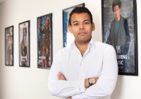 Jamie Njoku-Goodwin is stepping down as Chief Executive Officer at UK Music. He is photographed in UK Music Offices against a row of photos on the wall. He is wearing a white shirt, folding his arms, and looking at the camera.