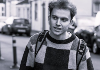 Black & white image of Archie Rowell, Activate Trainee Producer. He is a young, white man. He is photographed outside on a road with parked cars. He is looking to the side, with his mouth slightly open. He is wearing a jumper and backpack.