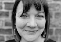Emily Bowman, incoming Managing Director, Junction Arts. A black-and-white, close-up image of Bowman. There is a blurred, brick wall in the background - Bowman is the focus of the image. She has dark hair and short bangs. She is smiling at the camera.