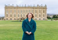 Jane Marriott is wearing a long, fur blue coat. She has ginger hair and is smiling at the camera. She is stood on the lawn in front of Chatsworth House.