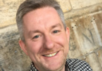 Gordon Morrison, incoming Chief Executive Officer (CEO) of Association for Cultural Enterprises. He is a white man standing against a brick wall. He has grey hair, stubble and is smiling at the camera. He wears a tartan shirt.