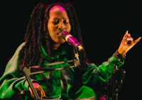 A black woman with elbow length long twists wearing a green outfit sitting down speaking into a microphone.