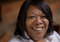 Stella Kanu, Shakespeare's Globe incoming CEO. Kanu is a Black woman with short, dark brown hair; she sits in the Globe, though the background is blurred. She smiles at the camera.