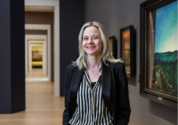 Karin Hindsbo standing in an art gallery. She has blonde hair, wears a black blazer and black/white striped shirt.