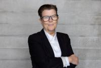 Kash Bennett, incoming President, Society of London Theatre. She is a white woman wearing large, black glasses. She wears a black blazer and white shirt, and has short, styled brown hair. There is a grey brick wall behind her.