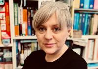 Alison McCrudden has short silver hair with green eyes. She is looking into the camera. There are books in the background.