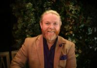 Michael Beavan, the Deputy CEO of PGGB, is sitting on a chair in front of a bush of ivy. He is a white man with a ginger hair and beard, wearing a mustard-colored blazer with a black shirt underneath. The photo appears to have been taken in the evening or at night. He is smiling at the camera.