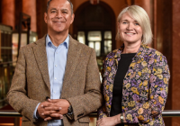 (L to R) Armoghan Mohammed (Chair), Kate Vokes (Deputy Chair), Royal Exchange Theatre. They stand in the theatre together. Mohammed is an Asian man, wearing a tweed brown blazer and blue shirt. Vokes is a white women with white-blonde hair. She wears a black shirt/dress, with a flower-patterned blazer.