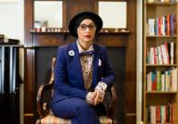 Amerah Saleh, Apple and Snakes Chair of the Board, a Western Asian woman, sits on a chair in an open room. She has her legs crossed and is looking at the camera, not smiling. Saleh wears red lipstick, black glasses, a top hat and hijab, along with a navy blue suit with a patterned white & black shirt.