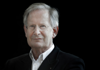 Sir John Eliot Gardiner, Principal Guest Conductor Emeritus, Philharmonia Orchestra. He is a white man with grey hair wearing glasses. He wears a black and white suit, and stands in front of a black background.