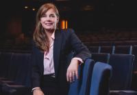 Theatre Royal Plymouth Chair of the Board Dame Darcey Bussell. She is seated in a theatre auditorium. She rests her elbow on the adjacent chair, smiling at the camera. She wears a dark blue suit with a pink shirt. Her hair is light brown, styled to the side.