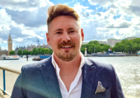 Tom O'Connell, incoming Executive Producer, DLAP Group. He is a white man with light hair, and a moustache/beard. He wears a navy suit with a white shirt, and stands on a bridge on the Thames in central London. It is a sunny day, and you can see Big Ben and boats in the background.