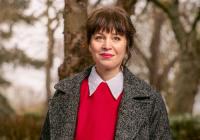 Kate Wasserberg, Artistic Director Theatr Clwyd. She is a white woman, stood outside with trees in the background. She is wearing red lipstick, a red jumper, white shirt and grey coat. She has brown hair with a full fringe and wears her hair up.