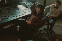 A women sits by a music recording desk, with a man sat behind her. both of them have sad expressions