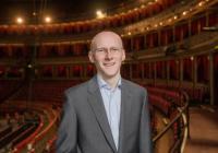 James Ainsclough smiling at the camera with the Royal Albert Hall auditorium in the background. He is bald, wears glasses and is wearing a suit.