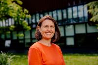 Kathy Bourne, Executive Director of Chichester Festival Theatre. She has short, brown hair and wears a bright orange top. She stands outside on grass, there are trees and a large black building behind her with many windows. She wears a large smile, and is looking at the camera.